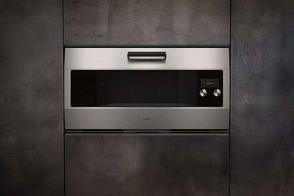 A stainless steel Gaggenau built-in microwave oven set into a dark gray, textured wall. The oven door is closed, and two control knobs are visible to the right of the display.