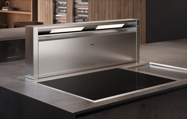 A stainless steel Gaggenau downdraft extractor hood integrated into a kitchen island. The hood is raised above a black induction cooktop, with subtle lighting illuminating the cooking surface.