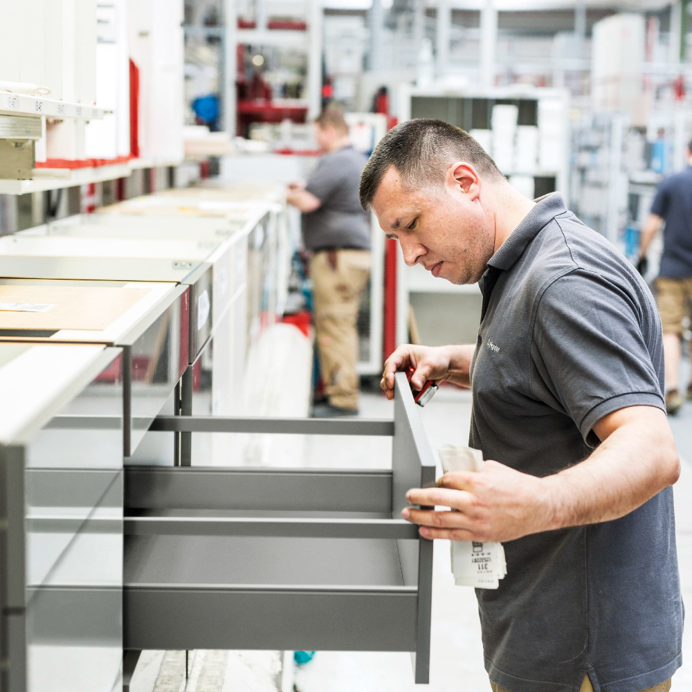 A worker inspects a gray kitchen drawer during the assembly process in a factory setting.