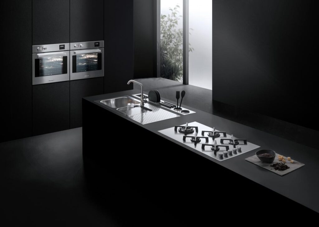 Minimalist black kitchen with built-in Barazza double ovens, a sink, and a gas cooktop on the island.