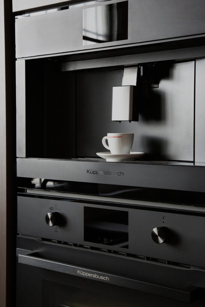 A built-in Kuppersbusch black coffee machine dispensing coffee into a white cup placed on a small shelf. The coffee machine is situated above a Küppersbusch oven.