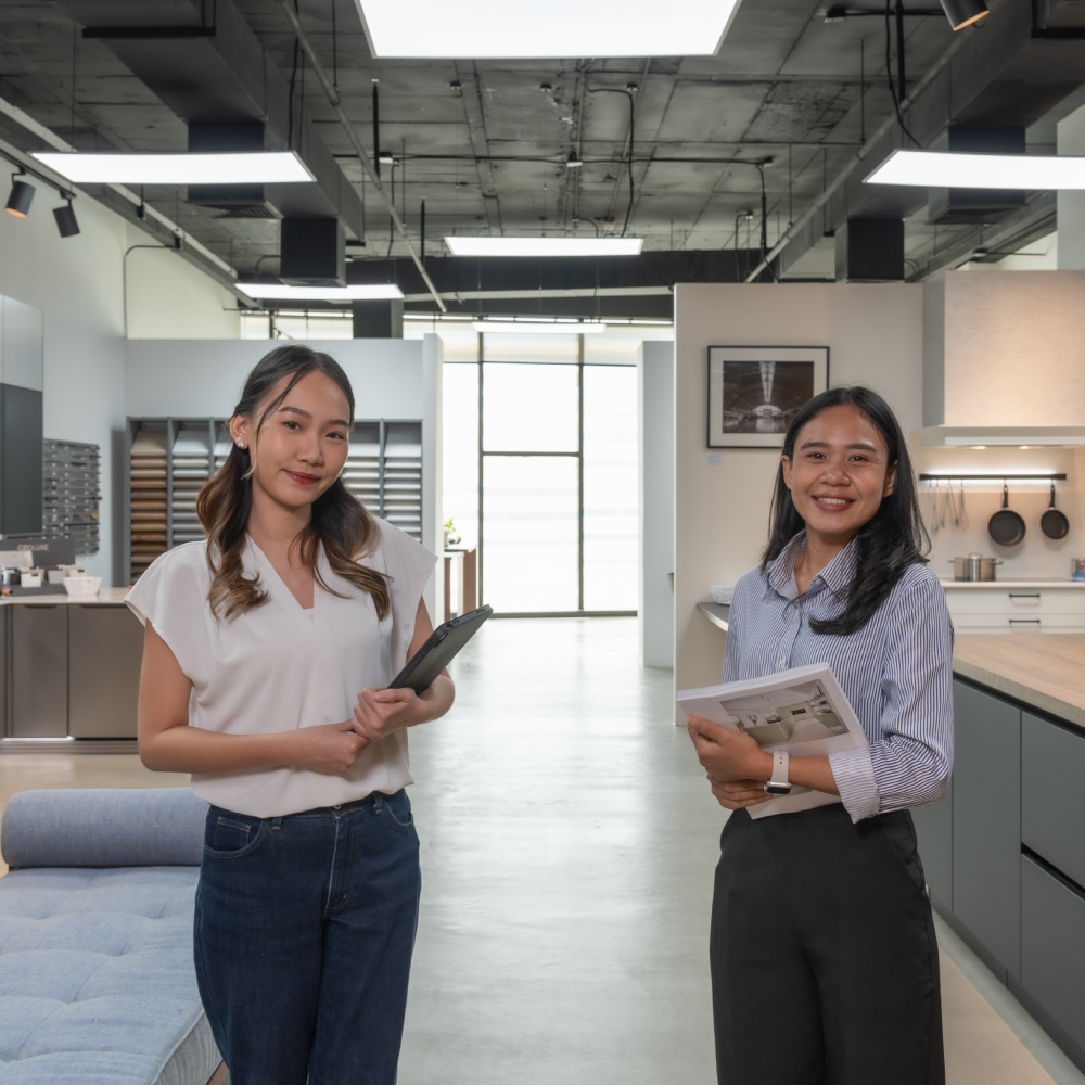 Two German Kitchen Design staff stand in a German Kitchen Design's showroom in River City Bangkok. One holds a tablet and the other a brochure.