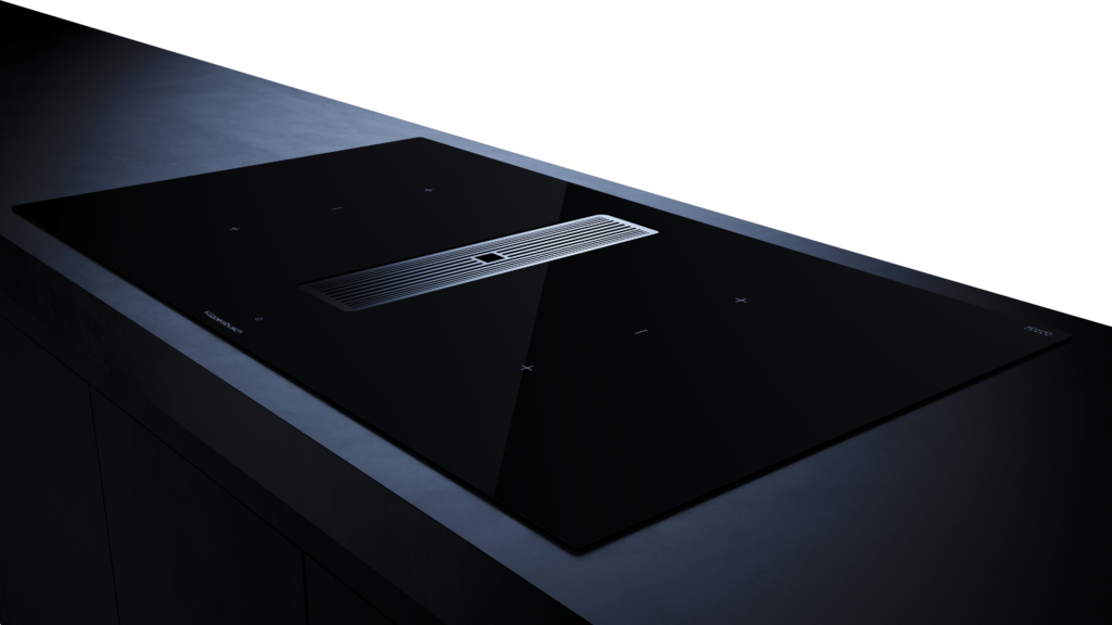 A black glass Kupperbusch induction cooktop with an integrated downdraft ventilation system in the center. The sleek cooktop is set into a concrete-like countertop, with touch controls indicated by small crosses.