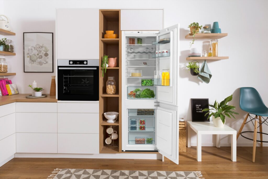 White kitchen with an open Gorenje refrigerator showing organized food, an oven, and open shelving.