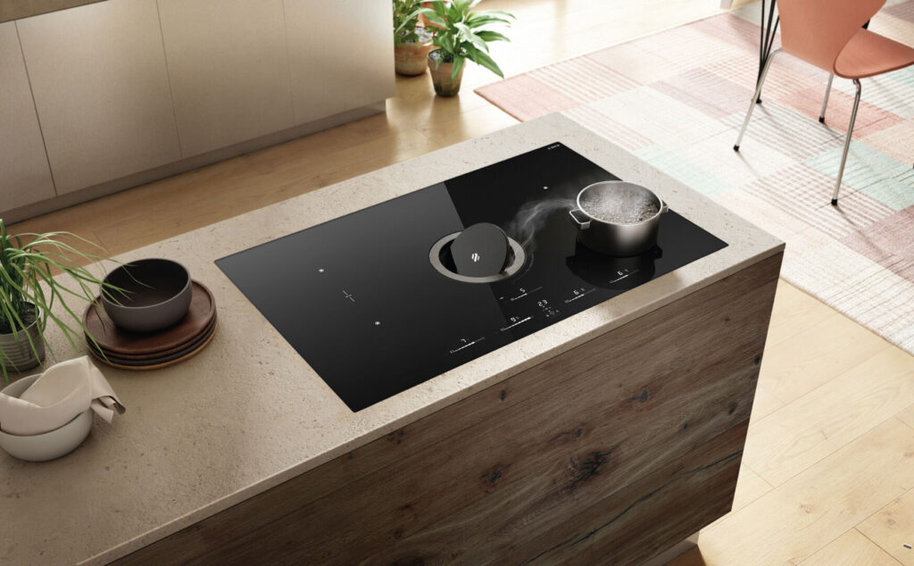 Black Elica induction cooktop with integrated Elica downdraft ventilation on a kitchen island with a mixed material countertop of concrete and wood. A pot of boiling water sits on the cooktop.